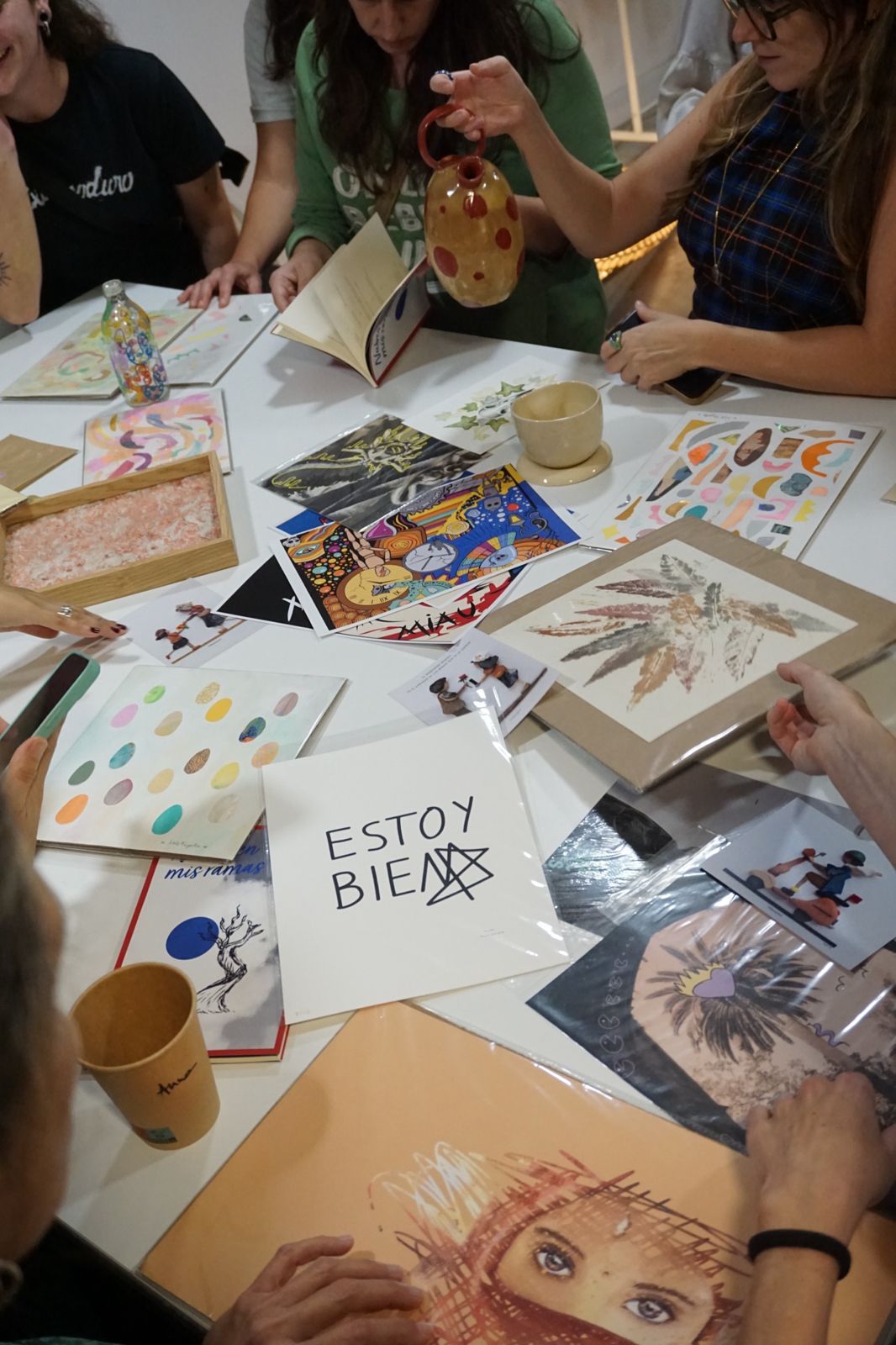 Mesa llena de obras durante un intercambio Arte x Arte: ilustraciones, cerámica, libros y láminas