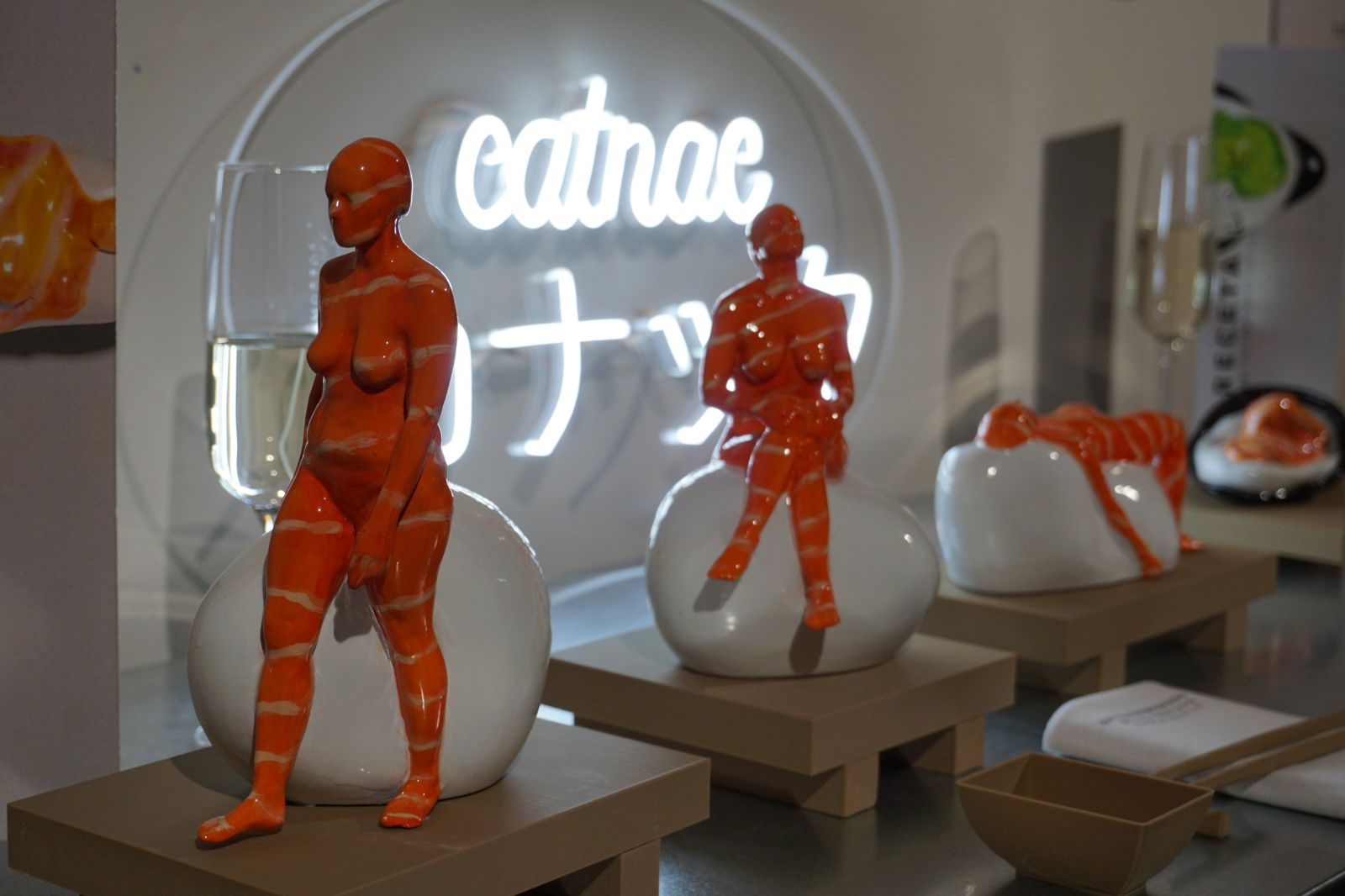 Esculturas de figuras naranjas sobre bases blancas, con un rótulo de neón, en una edición de Arts & Brands