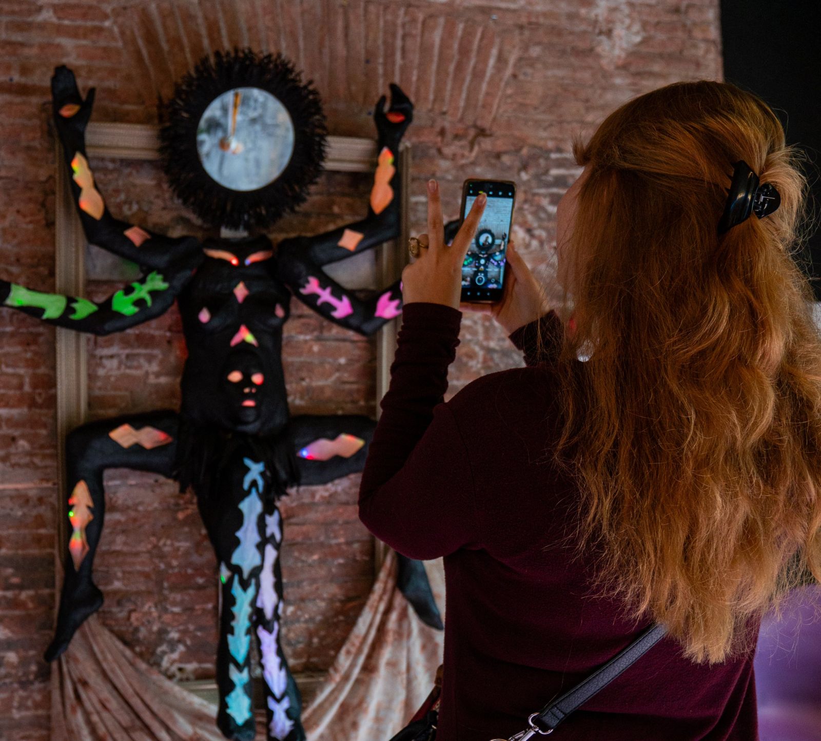 Una visitante fotografía con el móvil una escultura textil de colores colgada en una pared de ladrillo